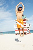 A young man with curly hair and a beard wearing bathing shorts jumping on the beach