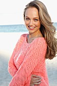 A young blonde woman by the sea wearing a pink summer jumper