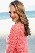 A young blonde woman by the sea wearing a pink summer jumper