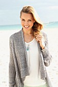 A blonde woman on a beach wearing a white blouse and a grey cardigan