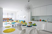 White, open-plan interior; dining area with classic shell chairs opposite white fitted kitchen with round, yellow rugs providing a splash of colour