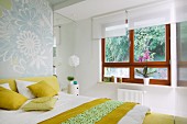 Bedroom with double bed, white and yellow bed linen and scatter cushions and wooden windows with white roller blinds and view of garden