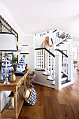 Woman walking down staircase with white balustrade in elegant interior; table lamps on console table to one side