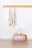 Gebogener Holzhocker mit gerollten Handtüchern vor Wand mit Wandhakenleiste und aufgehängten Handtüchern