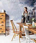 Young woman next to rustic wooden table, wooden chair, white-painted chair and chest of drawers in front of mural of grey cloudy sky