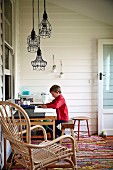 Kind am Schreibtisch und verschiedene Pendelleuchten mit Drahtlampenschirmen, im Vordergrund Rattanarmlehnsessel