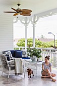 Frau und Hund auf überdachter Veranda mit Stuhl und Bank um Couchtisch aus weißem Rattan, vor verziertem Geländer mit geschnitzten Holzstützen