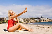 A platinum blonde woman on a beach wearing a red bathing suit taking a selfie