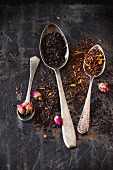 Dried black tea and rooibos with rose buds in vintage spoons on a black metal surface
