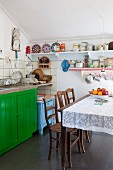 Esstisch mit Spitzendecke und antike Stühle, dahinter grün lackierter Unterschrank mit Spülbecken in schlichter Küche