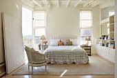 Stuhl im Rokoko Stil vor Doppelbett mit Rüschen Tagesdecke in hellem Schlafzimmer, weiße Holzdecke