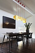 Designer dining table with retro character and matching black chairs below row of pendant lamps with pale yellow, paper lampshades