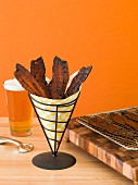 Candied bacon strips in a paper cone with more on a wire rack