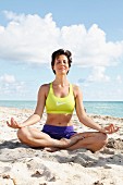 A woman with short, brown hair meditating on the beach wearing a sports bra and shorts