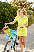 Junge, blonde Frau in gelbem Sommerkleid schiebt ein Fahrrad