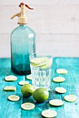 A glass of water with lime slices with a blue siphon bottle in the background