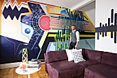 Artist in front of creative, graphic mural; aubergine corner sofa and vintage side table