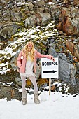 Blonde Frau in Minikleid mit breitem Federvolant, Daunenjacke und Strumpfhose in den Bergen vor 'Nordpol' Schild
