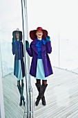 Lachende Frau mit großem Hut, Rock, lila Kurzmantel und Stiefeln an Fensterfront