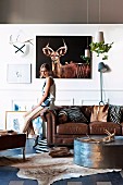 Safari-style lounge - animal skins and hunting trophies on floor, wall and coffee table and young woman perched on arm of brown leather sofa