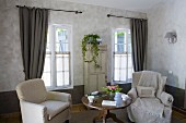 Two armchairs with loose covers flanking round Biedermeier table in front of windows with long curtains and potted ivy on old cabinet