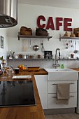 Wooden worksurfaces, white base units, cooking utensils on wall-mounted shelves and dark red lettering on wall in country-house-style kitchen