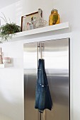 Cooks' apron hanging from handle of stainless steel fridge below white floating shelf