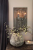 Vintage vase of flowering branches on table below lit candles on carved wooden candle scone