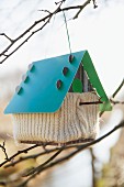A birds house with a knitted cover hanging on a twig