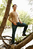 A young man standing on a log by a river wearing a shirt and jeans