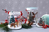 DIY Christmas arrangement of plastic animals and plastic fir trees in jam jars decorated with lace trim