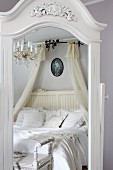 Mirror with white, carved wooden frame reflecting double bed with white bed linen and canopy