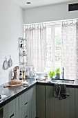 Kitchen with dark worksurface on L-shaped counter and integrated sink below window with crocheted curtains