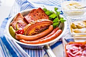 Wiener Würstchen, Bauchspeck und Leberkäse in einer Pfanne; dazu Ketchup, Senf und Meerettich