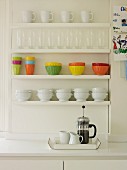 White and coloured crockery on white-painted, wall-mounted shelves above cafetiere on tray on worksurface