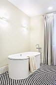 Free-standing designer bathtub and floor-mounted taps in elegant bathroom with floor-length curtains and black and white mosaic floor