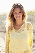 A young blonde woman on a beach wearing a white top and a pastel-yellow openwork jumper
