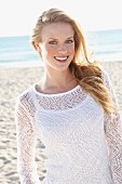 A young blonde woman on a beach wearing a white top and a transparent knitted jumper