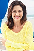 A brunette woman on a beach with her arms folded wearing a yellow knitted jumper