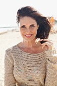 A brunette woman on a beach with her her blowing in the wind wearing a beige, openwork jumper