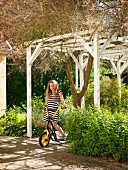 Girl on scooter under white pergola in summer garden