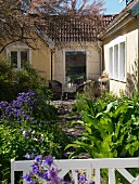 Terrassenplatz mit Korbmöbeln vor Hausfassade mit blühender Akelei im sommerlichen Garten