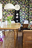 Wooden dining table below pendant lamp with white lampshade; long-case clock against leaf-patterned wallpaper in background