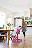 Woman walking past classic shell chairs and pink Tripp Trapp chair at wooden table in modern kitchen
