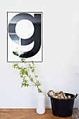 Framed, typographic poster of letter G on wall above leafy branch in white vase and bucket of logs on floor