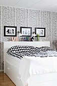 White box-spring bed with headboard and black and white patterned bed linen below framed family photos on patterned wallpaper