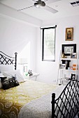 Yellow floral bedspread on black metal bed in front of small ladder-style shelves of ornaments