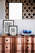 Coral under glass cover, framed black and white wallpaper patterns and blue glass bowl on copper chest of drawers below mirror reflecting wallpaper with geometric pattern