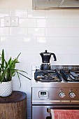 Edelstahl-Gasherd mit Espressokanne vor weißen Wandfliesen und Grünpflanze auf Baumstammhocker
