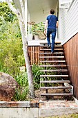 Man walking up rustic wooden steps without handrail into house entrance from green front garden planted with grasses and bushes